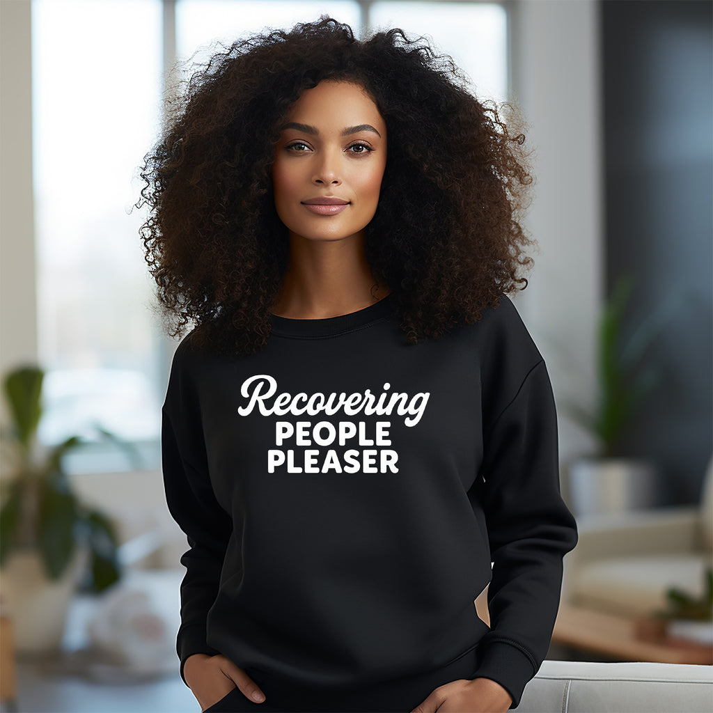 Woman wearing a black sweatshirt with 'Recovering People Pleaser' text in a blurred indoor setting